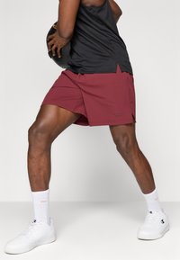 Bordové športové šortky s hladkou textúrou, s čiernym logom, bočnými rozparkami a kombinované s bielymi teniskami a ponožkami.