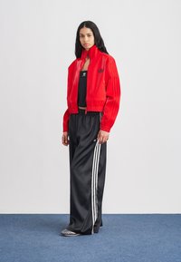 Kobieta ubrana w czerwoną kurtkę zapinaną na zamek, czarny top Adidas i czarne szerokie spodnie dresowe z białymi paskami, stojąca na niebieskim wykładzinie.