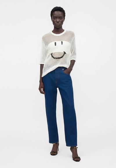 Γυναίκα φοράει λευκό πλεκτό πουλόβερ με μεγάλο μαύρο χαμογελαστό πρόσωπο, μπλε τζιν ψηλόμεσο και μαύρες ανοιχτές γόβες, στέκεται με το ένα χέρι στην τσέπη.