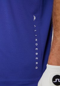 Blue athletic shirt with a smooth texture, featuring white vertical text "J. LINDEBERG" on the side. Close-up view of fabric and design.
