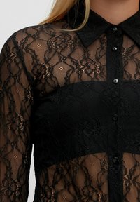 Zwarte bloemenkant blouse met lange mouwen, kraag en knopen, van doorzichtig materiaal dat de zwarte onderkleding eronder onthult.