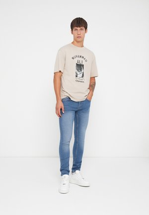 Camiseta de algodón beige con gráfico negro de Muhammad Ali, combinada con jeans ajustados azules y zapatillas blancas. Diseño simple, look casual.