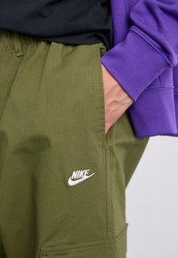 Pantaloni cargo Nike de culoare verde măslin, dintr-un material texturat, cu un logo alb și buzunar lateral. Asortați cu un hanorac violet.