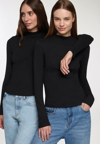 Dwie modelki noszą dopasowane czarne bluzki z długim rękawem i wysokimi kołnierzami, zestawione z jasnoniebieskimi dżinsami. Bluzki mają strukturalny, prążkowany wzór.