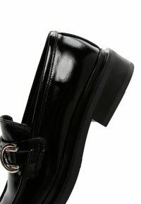 Black patent leather loafer with a rounded toe, low block heel, and a prominent silver buckle accent on a side strap. Smooth texture.