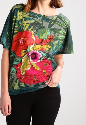 Femme portant une chemise à manches courtes verte avec un grand imprimé floral rouge, rose et jaune, et un pantalon noir, la main posée sur la hanche.