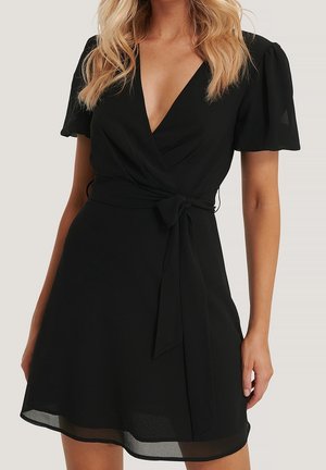 Femme portant une courte robe portefeuille noire avec des manches bouffantes et une ceinture nouée à la taille, montrant des cheveux blonds et des bras nus.