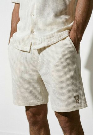 Uomo che indossa pantaloncini bianchi con texture a nido d'ape e camicia abbottonata coordinata, mani nelle tasche, logo dell'orso visibile sulla gamba sinistra dei pantaloncini.