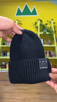 Zwarte gebreide muts met geribbelde textuur, met een medium hoogte en een zwart label met "C P H STUDIOS" in witte tekst. Gehouden door een hand.