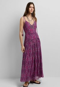Mujer con vestido morado sin mangas de largo hasta el suelo con estampado, tirantes con lazos, collar plateado en capas y sandalias marrones sobre un fondo gris.
