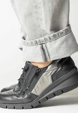 Zapatillas deportivas de cuero negro y metal gris con un acabado texturizado, que cuentan con cierre de cremallera y una suela de goma gruesa, combinadas con jeans con los puños doblados.