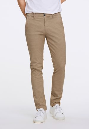 Uomo che indossa pantaloni beige slim fit con passanti per cintura e sneaker bianche, in piedi davanti a uno sfondo semplice.