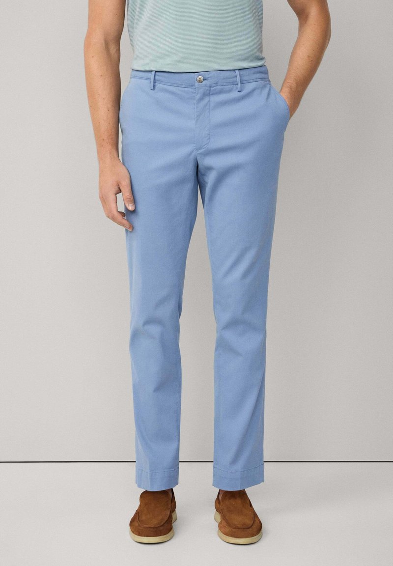 Light blue cotton trousers with a straight fit, front pockets, and a button closure. Worn with brown suede shoes. Minimalist design.