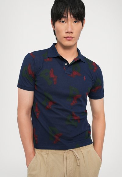 Ναυτικό πόλο μπλουζάκι με επαναλαμβανόμενο μοτίβο από κόκκινες φτερούγες και πράσινα γράμματα. Υλικό από μίγμα βαμβακιού με κλασικό γιακά και κοντά μανίκια.