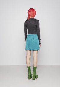 Schwarzes Glitzer-Turtleneck-Oberteil, teal-farbener Wildleder-A-Linien-Minirock und grüne Wildleder-Knielange Blockabsatzstiefel. Minimalistischer Hintergrund.