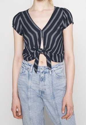 Top corto a righe navy con maniche corte, scollatura a V e dettaglio con nodo davanti, realizzato in Tessuto leggero, abbinato a jeans azzurri a vita alta.