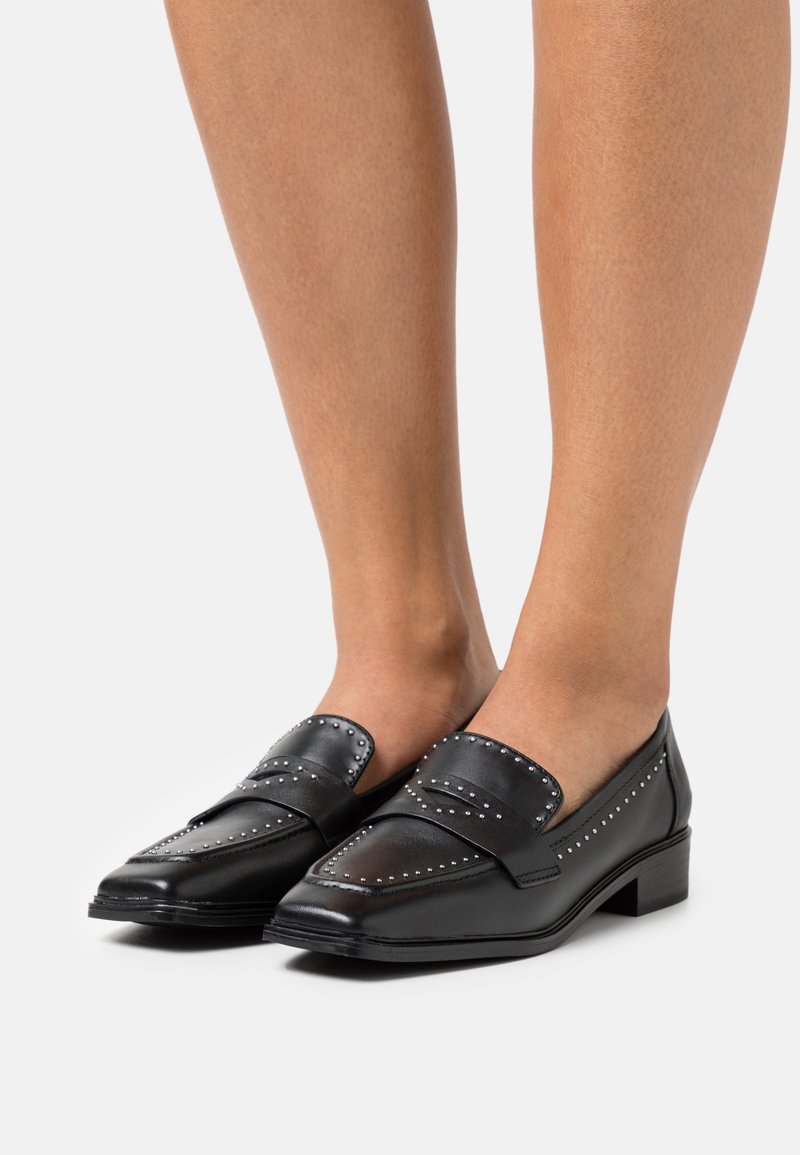 Mocassins en cuir noir avec des clous décoratifs sur le dessus, petit talon carré et bout arrondi, présentant une texture lisse et un design élégant.