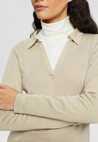 Top beige a maniche lunghe con scollo a V e colletto, realizzato in tessuto morbido. Presenta una silhouette aderente e una texture liscia. Sotto, una maglietta a collo alto bianca.