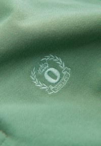 Tissu vert avec un petit emblème brodé comprenant un dessin circulaire, une couronne de feuilles et une couronne au-dessus du cercle.