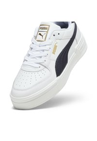Baskets blanches Puma avec bande latérale noire, logo Puma doré sur la languette et le côté, lacets blancs et zone perforée à l’avant.
