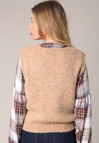 Pull en tricot sans manches beige avec une texture côtelée et un col rond, porté par-dessus une chemise à manches longues à carreaux. Accent argenté à l'arrière.