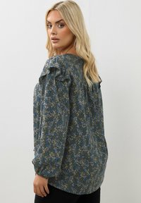 Blommig blus med ett blått och grönt mönster, volangiga axlar och långa ärmar, tillverkad av lätt tyg med en avslappnad passform.