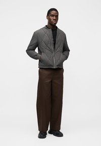 Chaqueta gris acolchada con cierre de cremallera y dos bolsillos laterales, combinada con pantalones marrones holgados y zapatillas negras.