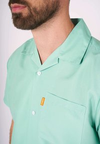 Minttu vihreä puolihihainen paita, joka on valmistettu sileästä kankaasta. Paidassa on kaulus, napit ja etutasku, jossa on oranssi etiketti.