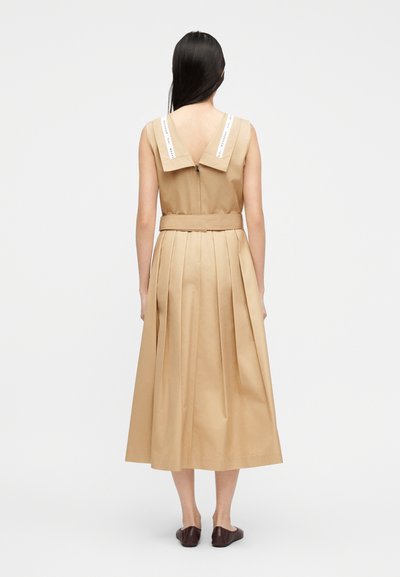 Γυναίκα με την πλάτη προς τον θεατή, φοράει μπεζ midi φόρεμα χωρίς μανίκια με φαρδύ γιακά και πλισέ φούστα, σε συνδυασμό με σκούρα καφέ μπαλαρίνες.