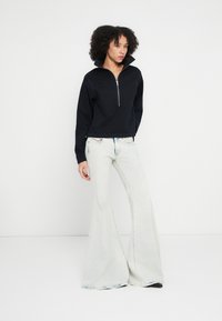 Μαύρο φούτερ με φερμουάρ μέχρι τον λαιμό και ψηλό γιακά, συνδυασμένο με ανοιχτό μπλε καμπάνα jeans. Το σύνολο έχει άνετη εφαρμογή και απαλή υφή υφάσματος.