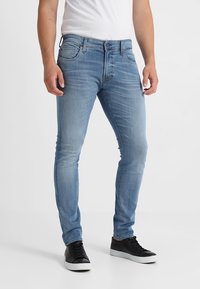 Jeans de mezclilla azul claro con un corte ajustado, que cuentan con cinco bolsillos, cierre de botón y un ligero desvanecimiento en la tela. Llevados con zapatillas negras.