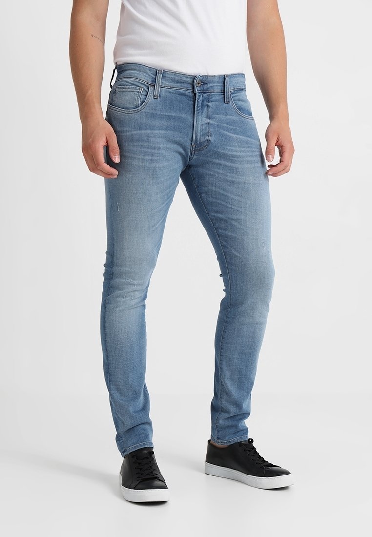 Jeans de mezclilla azul claro con un corte ajustado, que cuentan con cinco bolsillos, cierre de botón y un ligero desvanecimiento en la tela. Llevados con zapatillas negras.