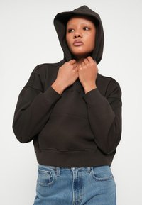 Sweatshirt castanho cropped com capuz em material suave, com uma costura à frente e detalhe de logo subtil. Combinado com jeans de ganga azul.