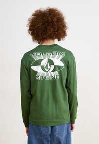 Grønn langermet t-skjorte med en stor hvit grafisk design på baksiden med teksten "VOLCOM" og "STONE". Laget av bomull, avslappet passform.