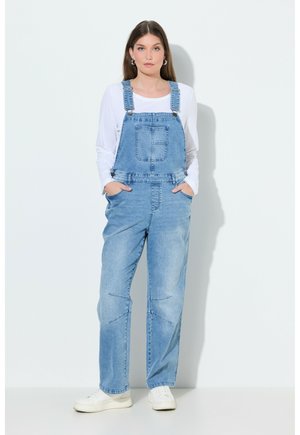 Jeune femme debout, les mains dans les poches, portant une salopette en jean bleu clair, un t-shirt à manches longues blanc et des baskets blanches, sur un fond uni.