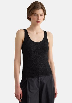 Mujer con cabello corto y castaño, llevando una camiseta sin mangas negra con textura y pantalones negros sueltos, de pie frente a un fondo liso.