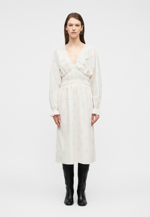 Kvinne iført en langermet, lys blomstermønstret midikjole med dyp V-hals og svarte knelange støvler, stående mot en hvit bakgrunn.