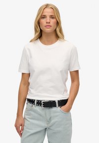 Hvid bomulds t-shirt med korte ærmer, rund halsudskæring og subtilt præget logo, parret med lyseblå jeans og et sort nittet bælte.
