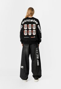 Sudadera negra con estampados gráficos de latas en la parte posterior, combinada con pantalones vaqueros oscuros de gran tamaño y zapatillas gruesas.