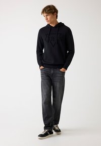 Felpa nera con la parola "GUESS" in un design triangolare, abbinata a jeans grigi e sneakers nere con dettagli color crema.