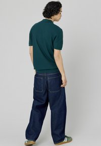 Polo shirt court à manches courtes de couleur vert foncé avec une finition texturée, associé à un jean en denim bleu foncé ample avec des coutures visibles et des poches arrière.