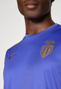 Modrá športová košeľa vyrobená z hladkej tkaniny, s zlatým logom AS Monaco FC a čiernym Nike swooshom na hrudi.
