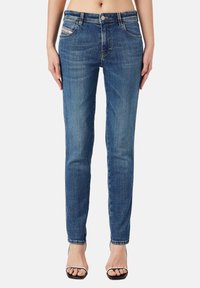 Mujer vestida con jeans azul de corte Slim-fit de tiro medio y tacones abiertos negros, de pie contra un fondo claro y liso.