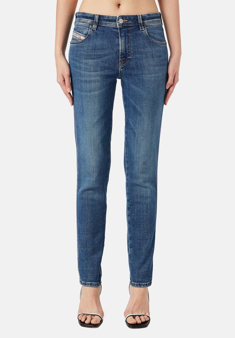 Mujer vestida con jeans azul de corte Slim-fit de tiro medio y tacones abiertos negros, de pie contra un fondo claro y liso.