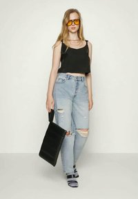 Mulher jovem vestindo um top cropped preto, jeans claros rasgados, óculos de sol com tonalidade laranja, segurando uma grande bolsa preta, de pé contra um fundo simples.