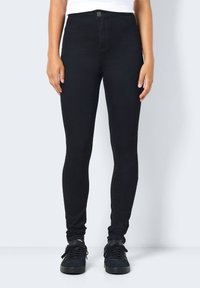 Jeans negros ajustados con un diseño ceñido, talle alto, cierre de botón y tela elástica. Combinados con zapatillas negras.