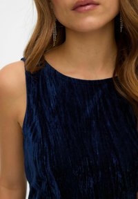 Navyblå sammets topp med en mjuk, texturerad yta, med breda axelremmar och rundad halsringning. Hängande örhängen synliga.