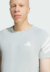 Camiseta deportiva de color gris claro con una superficie texturizada y rayas blancas en los hombros. Presenta un logo blanco en el lado izquierdo del pecho.