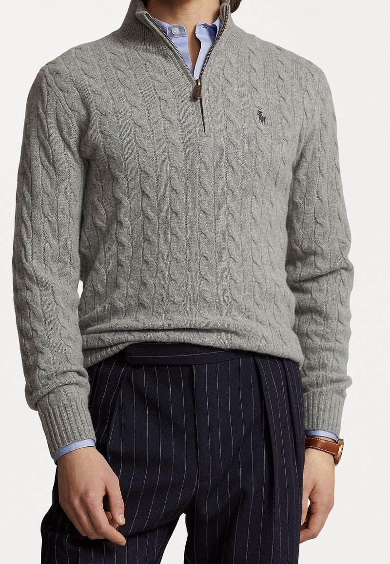 Homme portant un pull gris en maille torsadée avec un quart de zip, par-dessus une chemise bleue, un pantalon rayé bleu marine et une montre en cuir marron.
