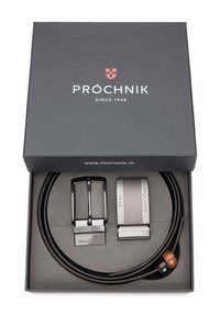 Brązowy skórzany pasek z dwoma klamrami: klasyczną czarną w stylu, oraz nowoczesną srebrną z rytowanymi detalami, umieszczony w czarnym pudełku.